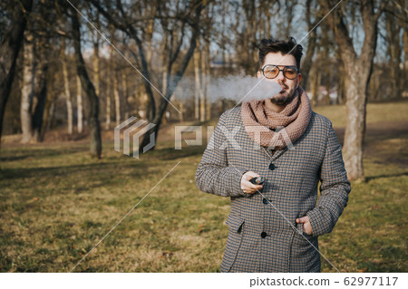 Confident young bearded male in sunglasses vape electronic cigarette. A young handsome bearded hipster man vaping e-cigarette outdoors in the park. Close up. 62977117