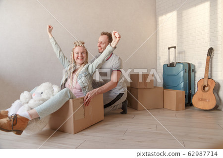 Happy young couple moving to a new apartment. A man pushes a woman sitting in a box with arms raised. Packed things in the background. Happy young couple moving to a new apartment. A man pushes a woman sitting in a box with arms raised. Packed things in the background. 62987714