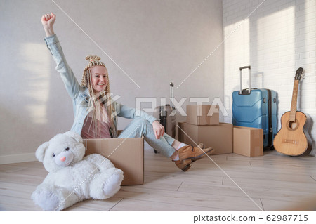 Young woman with pigtails is happy to move to new flat. Sits in a box with raised hand. Young woman with pigtails is happy to move to new flat. Sits in a box with raised hand. 62987715
