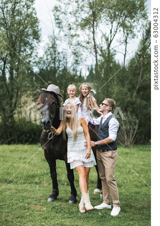 Happy family, parents and children, enjoying the horse presence. Girls are riding the horse, wearing hat. Man and woman parents having fun and laughing. Countryside, horse riding outdoors Happy family, parents and children, enjoying the horse presence. Girls are riding the horse, wearing hat. Man and woman parents having fun and laughing. Countryside, horse riding outdoors 63004312