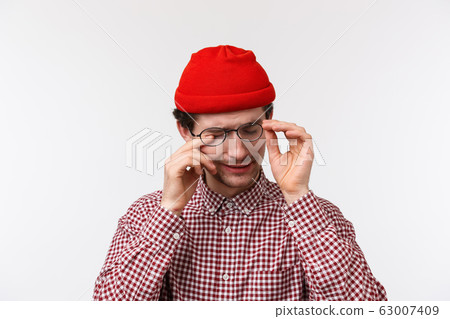Close-up portrait of handsome caucasian hipster man in red beanie, take-off glasses to scratch eye, feel tired after looking computer screen, programmer have itchy eye, standing white background 63007409
