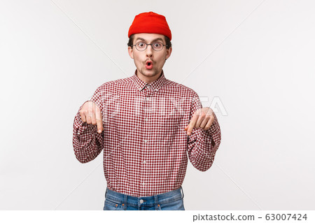Waist-up portrait excited and surprised amused handsome caucasian hipster in beanie and glasses telling someone what he found in online store, pointing fingers down tell to quickly take look 63007424
