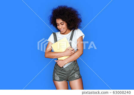Exam Preparation. Mulatto girl wearing denim overall standing isolated on blue with notebooks smiling joyful Exam Preparation. Mulatto girl wearing denim overall standing isolated on blue with notebooks smiling joyful 63008240