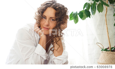 Portrait of a beautiful woman sitting at home with plants and smiling at camera Portrait of a beautiful woman sitting at home with plants and smiling at camera 63008978