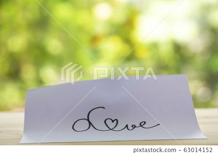 Hand writing " Love " on white paper texture, Wooden table, Bokeh garden background, Close up shot, Selective focus, Stationery concept 63014152