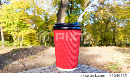 a red corrugated paper cup with coffee covered with a brown lid and a white drinking straw stands in the forest on a gray wooden table against the background of nature and trees. Copy space. 63040585
