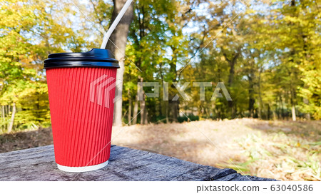 a red corrugated paper cup with coffee covered with a brown lid and a white drinking straw stands in the forest on a gray wooden table against the background of nature and trees. Copy space. a red corrugated paper cup with coffee covered with a brown lid and a white drinking straw stands in the forest on a gray wooden table against the background of nature and trees. Copy space. 63040586