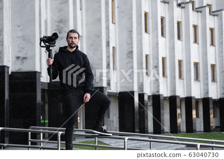 Young Professional videographer holding professional camera on 3-axis gimbal stabilizer. Pro equipment helps to make high quality video without shaking. Cameraman wearing black hoodie making a videos. 63040730