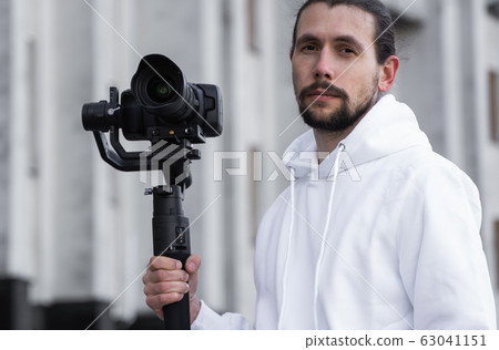 Young Professional videographer holding professional camera on 3-axis gimbal stabilizer. Pro equipment helps to make high quality video without shaking. Cameraman wearing white hoodie making a videos. Young Professional videographer holding professional camera on 3-axis gimbal stabilizer. Pro equipment helps to make high quality video without shaking. Cameraman wearing white hoodie making a videos. 63041151