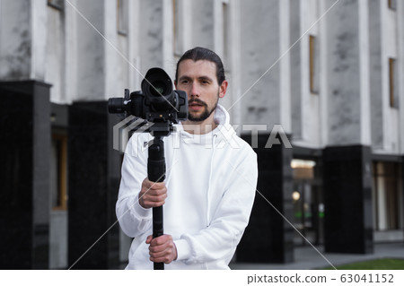 Young Professional videographer holding professional camera on 3-axis gimbal stabilizer. Pro equipment helps to make high quality video without shaking. Cameraman wearing white hoodie making a videos. Young Professional videographer holding professional camera on 3-axis gimbal stabilizer. Pro equipment helps to make high quality video without shaking. Cameraman wearing white hoodie making a videos. 63041152