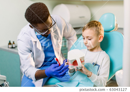 African-American ethnic black male dentist in blue latex gloves checking condition of teeth of little smiling boy. Little boy in blue dental chair in modern dentistry clinic African-American ethnic black male dentist in blue latex gloves checking condition of teeth of little smiling boy. Little boy in blue dental chair in modern dentistry clinic 63043127