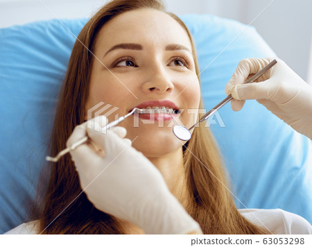 Smiling young woman with orthodontic brackets examined by dentist at dental clinic. Healthy teeth and medicine concept Smiling young woman with orthodontic brackets examined by dentist at dental clinic. Healthy teeth and medicine concept 63053298