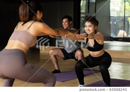 Group of happy Asian women and man doing squat exercises on yoga mats in aerobics class. Young sporty people smiling while working out in gym studio. 63088243