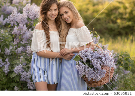 Two young girls dressed in beautiful clothes stand against the background of lilac bushes 63095931