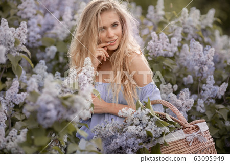 Charming blonde model against the backdrop of lilac bushes holds a straw basket with flowers in her hands 63095949