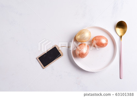 Greeting card with handmade painted bright eggs on a plate and golden spoon on a light grey marble background. 63101388