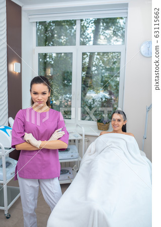 Dark-haired cosmetologist wearing uniform standing near client 63115662