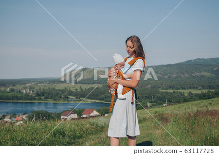 woman with baby in a loose dress with loose hair walks in a meadow. Village houses, forest and river as background. The concept of summer, warmth, freedom, village life, sunburn, childhood 63117728