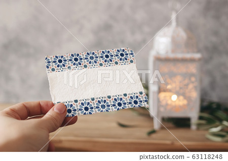 Closeup of hand holding blank paper card with watercolor Moroccan pattern. Greeting card mock-up scene. Ramadan Kareem design. Blurred background with White arabic lantern, olive leaves on table. Closeup of hand holding blank paper card with watercolor Moroccan pattern. Greeting card mock-up scene. Ramadan Kareem design. Blurred background with White arabic lantern, olive leaves on table. 63118248