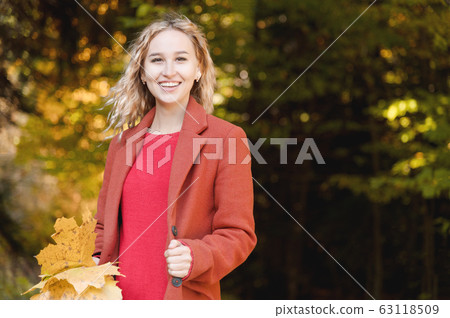 Portrait of attractive caucasian young blonde girl in red coat with a bouquet of fallen yellow leaves smiling in the autumn forest. The concept of autumn and fall holidays and weekends 63118509