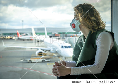 Woman with a mask on her mouth protects against the virus. She looks sad through the window at the airport on planes. Aerial connections canceled due to a coronavirus 63118597