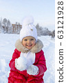 Little beautiful girl in a red coat and white scarf holds a round snowball in her hands and smiles. Snow-covered path and snowdrifts behind her. Children's entertainment during the winter holidays 63121928