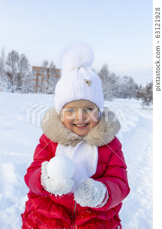 Little beautiful girl in a red coat and white scarf holds a round snowball in her hands and smiles. Snow-covered path and snowdrifts behind her. Children's entertainment during the winter holidays 63121928