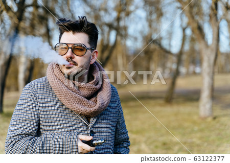 Confident young bearded male in sunglasses vape electronic cigarette. A young handsome bearded hipster man vaping e-cigarette outdoors in the park. Close up. Confident young bearded male in sunglasses vape electronic cigarette. A young handsome bearded hipster man vaping e-cigarette outdoors in the park. Close up. 63122377