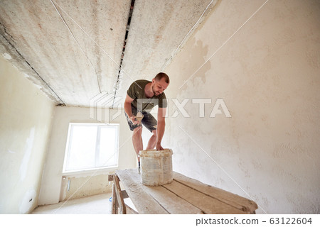 Male is making repairing in apartment by putting putty on the walls and standing on wooden stand against a window 63122604