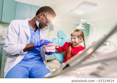 The child girl in dentist's chair having fun while brushing teeth with a male African American doctor 63124614
