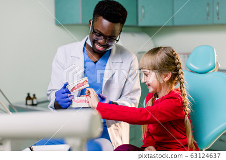 African dentist shows how to brush teeth to a small Caucasian girl a patient, using a artificial jaws. Dentistry, teeth hygiene concept 63124637