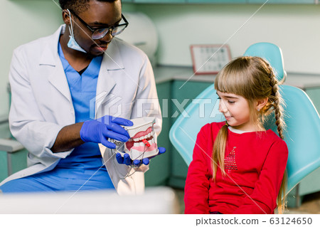 Little patient at the dentist's office. Young African American dentist talking with patient small girl holding artificial jaws in his hands. 63124650