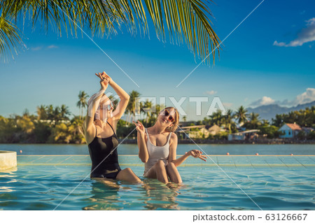 Two beautiful young woman sitting by the poolside of a resort swimming pool during summer holiday Two beautiful young woman sitting by the poolside of a resort swimming pool during summer holiday 63126667