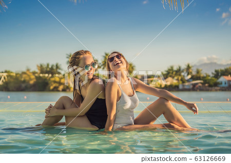 Two beautiful young woman sitting by the poolside of a resort swimming pool during summer holiday Two beautiful young woman sitting by the poolside of a resort swimming pool during summer holiday 63126669