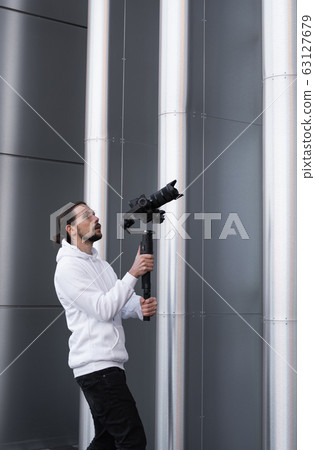 Young Professional videographer holding professional camera on 3-axis gimbal stabilizer. Pro equipment helps to make high quality video without shaking. Cameraman wearing white hoodie making a videos. 63127679