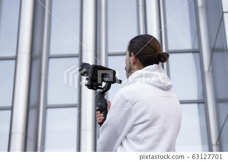 Young Professional videographer holding professional camera on 3-axis gimbal stabilizer. Pro equipment helps to make high quality video without shaking. Cameraman wearing white hoodie making a videos. 63127701