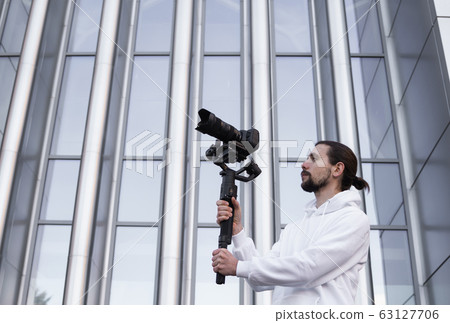 Young Professional videographer holding professional camera on 3-axis gimbal stabilizer. Pro equipment helps to make high quality video without shaking. Cameraman wearing white hoodie making a videos. 63127706