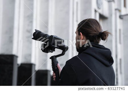 Young Professional videographer holding professional camera on 3-axis gimbal stabilizer. Pro equipment helps to make high quality video without shaking. Cameraman wearing black hoodie making a videos. 63127741
