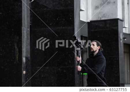 Young Professional videographer holding professional camera on 3-axis gimbal stabilizer. Pro equipment helps to make high quality video without shaking. Cameraman wearing black hoodie making a videos. Young Professional videographer holding professional camera on 3-axis gimbal stabilizer. Pro equipment helps to make high quality video without shaking. Cameraman wearing black hoodie making a videos. 63127755