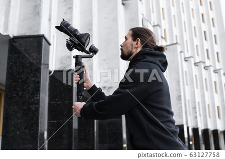 Young Professional videographer holding professional camera on 3-axis gimbal stabilizer. Pro equipment helps to make high quality video without shaking. Cameraman wearing black hoodie making a videos. Young Professional videographer holding professional camera on 3-axis gimbal stabilizer. Pro equipment helps to make high quality video without shaking. Cameraman wearing black hoodie making a videos. 63127758