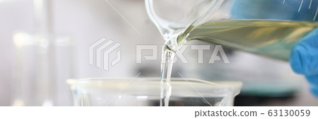 Scientist arm wearing blue protective gloves pouring yellow liquid at laboratory flask 63130059