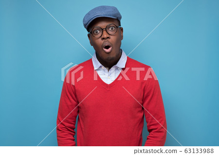 Black african american man in red sweater and blue hat looking surprised. 63133988