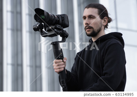 Young Professional videographer holding professional camera on 3-axis gimbal stabilizer. Pro equipment helps to make high quality video without shaking. Cameraman wearing white hoodie making a videos. 63134995