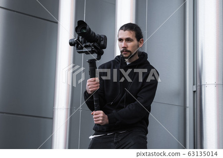 Young Professional videographer holding professional camera on 3-axis gimbal stabilizer. Pro equipment helps to make high quality video without shaking. Cameraman wearing white hoodie making a videos. 63135014