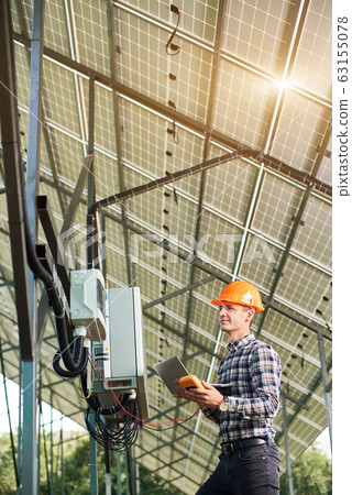 Checking the operation of solar power plant. Man in helmet and with laptop at station 63155078