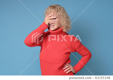 Senior woman covering face with hands and peeping through her fingers at camera. 63155146