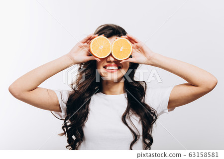 Young Brunette Woman Covering Eyes With Halves of Orange 63158581