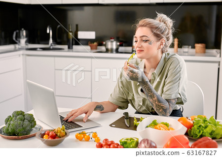 Leisure at Home. Young tattooed woman standing at kitchen\
watching video on laptop eating vegetable laughing cheerful while\
cooking