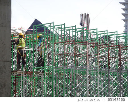 Construction workers wearing safety gear and safety harness while installing scaffolding at a high level in the construction site. 63168660