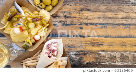 Tapas, pinchos, and wine, overhead panoramic shot on a wooden table. Cheese, jamon and olives, gildas with peppers and potato chips in an outdoors cafe with a place for text 63186409